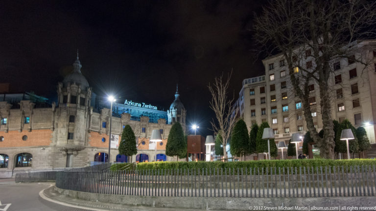 Azkuna Zentroa at night seen behind clipped trees, bare saplings, railings, and landscaped public space.
