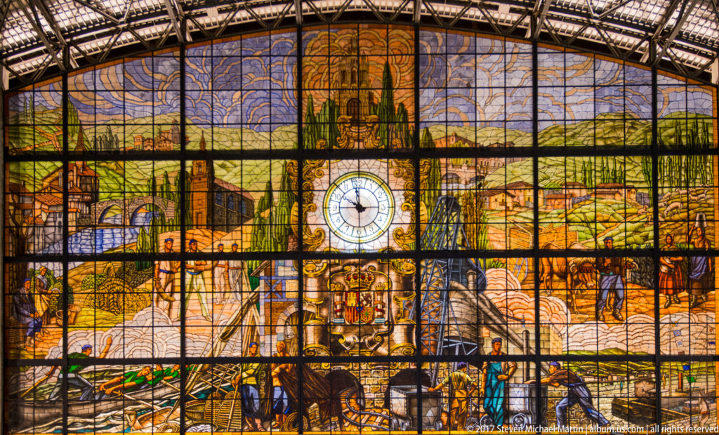 Close view of Bilbao-Abando station’s stained-glass mural with industrial and rural scenes, figures, central clock, and heraldic shield.