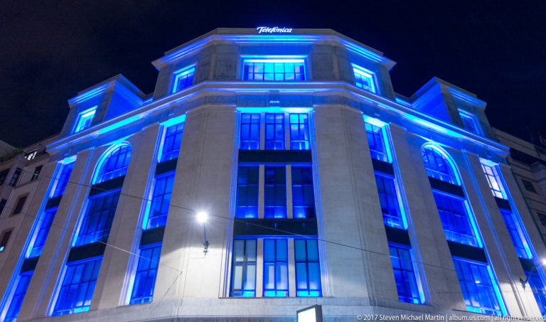 Low-angle night view of a Telefónica building outlined in bright blue light, with tall windows and the company name on the roofline.