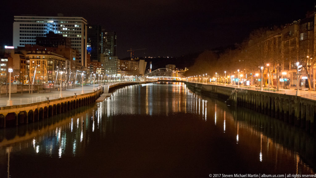 Long nighttime view down the Nervión estuary between illuminated Bilbao quays, with city buildings and reflections on both banks.