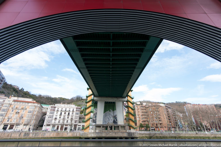 Underside of La Salve Bridge spanning the river, with apartment blocks visible across the opposite bank.