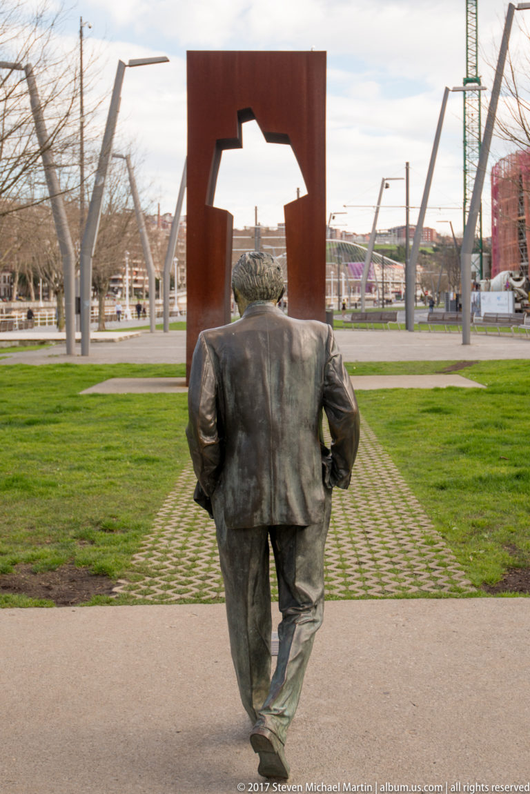 Bronze walking figure viewed from behind, aligned with a tall rusted-steel portal in a grassy urban park.