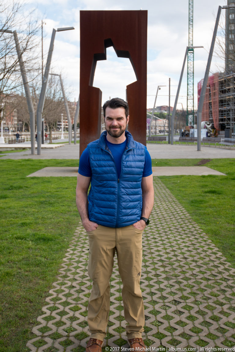 Man in blue vest standing beneath the opening of a large rusted-steel sculpture in a Bilbao park.