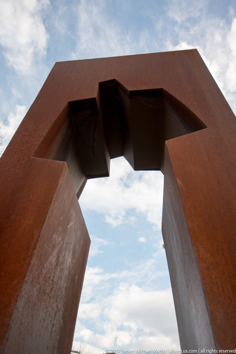 Low-angle view through a massive rusted-steel geometric portal, with its stepped opening silhouetted against the sky.