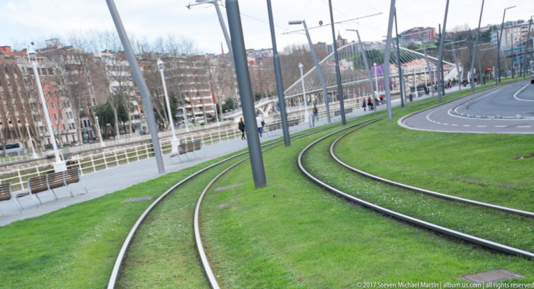 Curving tram tracks embedded in green grass beside a Bilbao boulevard with overhead poles and apartment buildings.