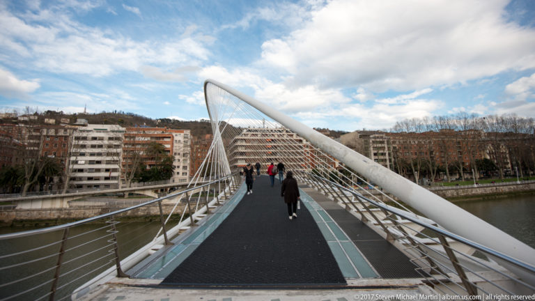 Pedestrians walking across a white cable-supported arched footbridge over the Nervión River in Bilbao.