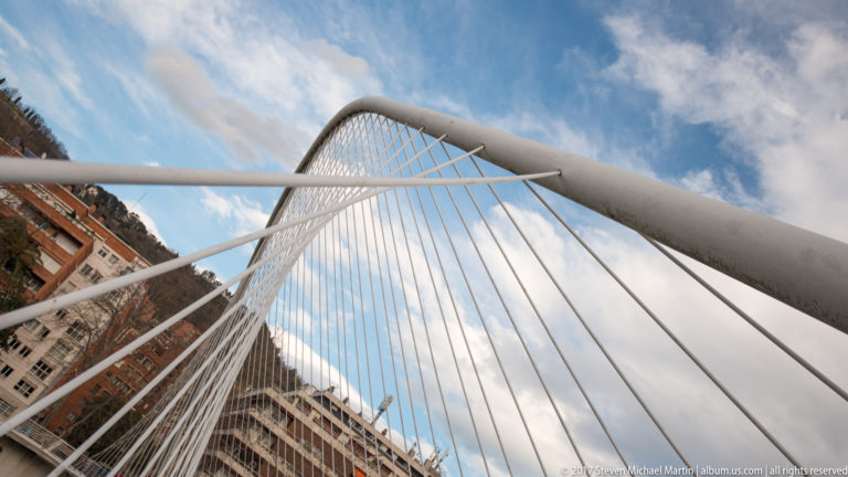 Upward view of a white pedestrian-bridge arch with closely spaced suspension cables against a blue, clouded sky.