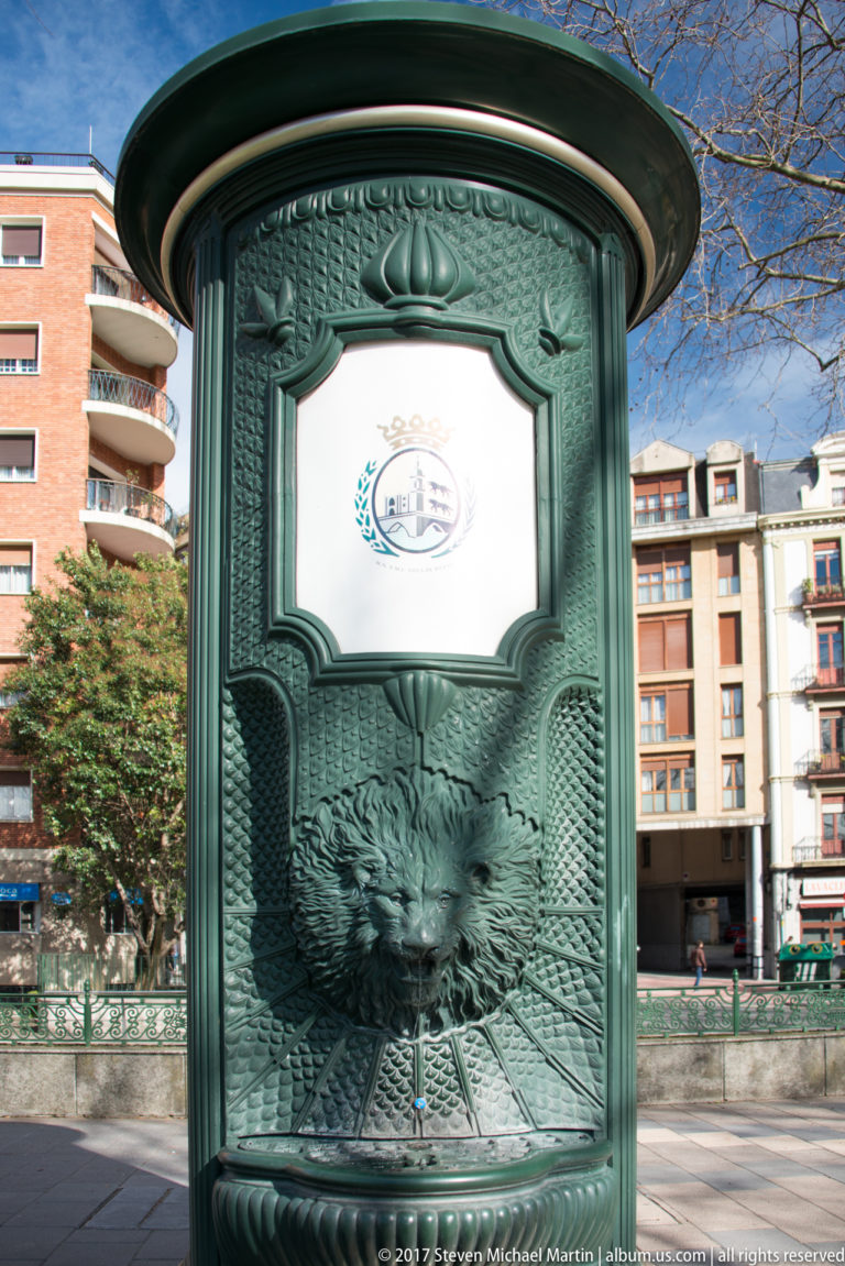 Dark-green ornamental metal information kiosk with relief eagle, set against apartment buildings in Bilbao.