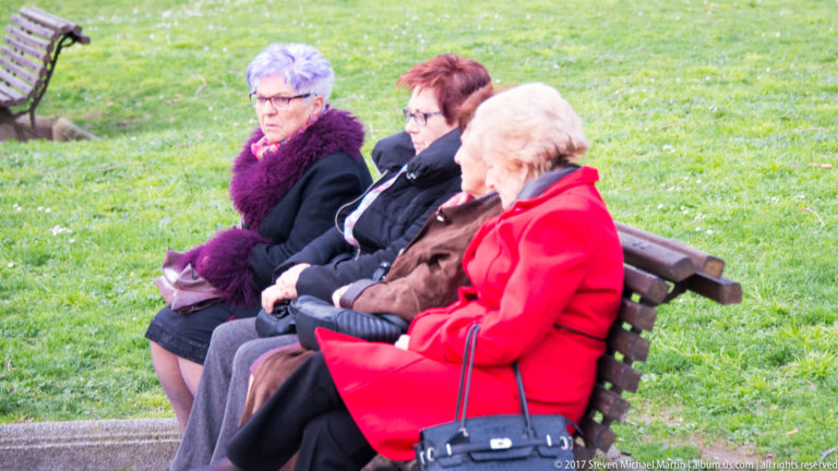 Three elderly women in winter coats sitting together on a wooden bench beside a green lawn.