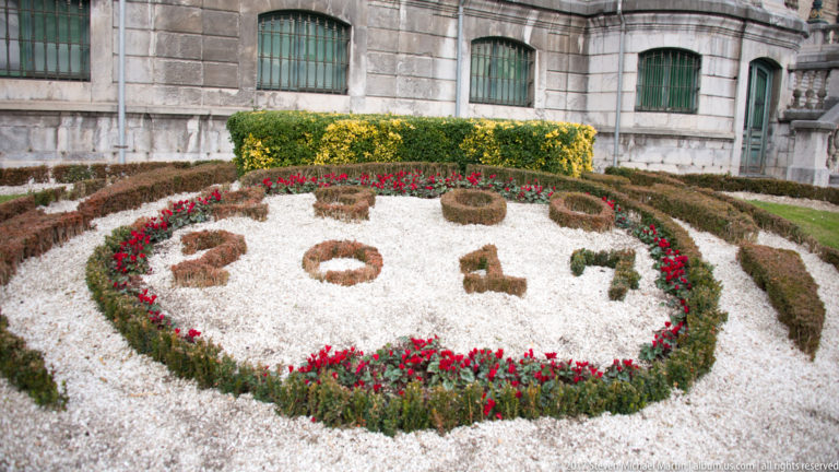 Formal flowerbed spelling 2017 in dark plants on white gravel, bordered by red flowers beside a stone building.