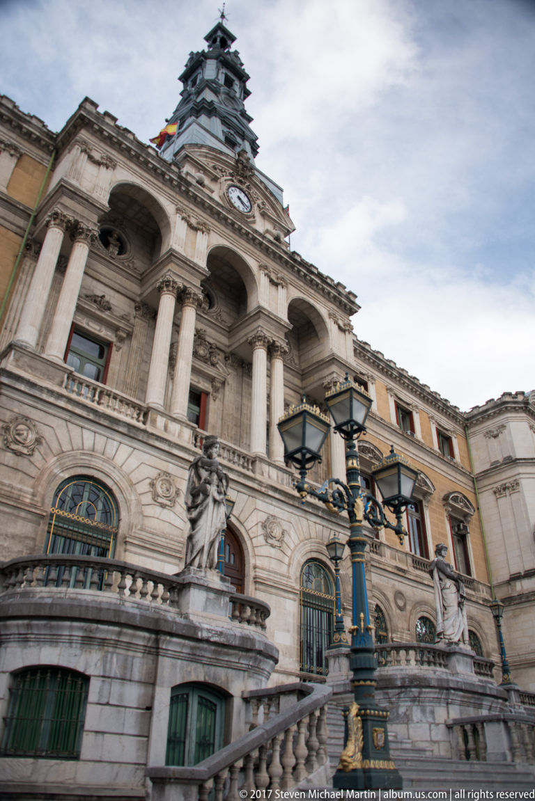 Ornate Bilbao City Hall façade with columns, arched windows, sculptural decoration, clock tower, and historic street lamps.
