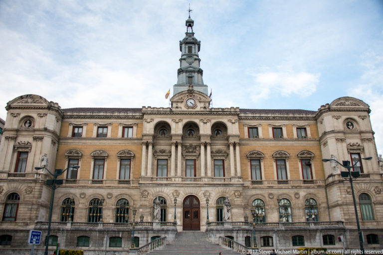 Frontal view of Bilbao City Hall with symmetrical wings, central arches, clock tower, and broad entrance steps.