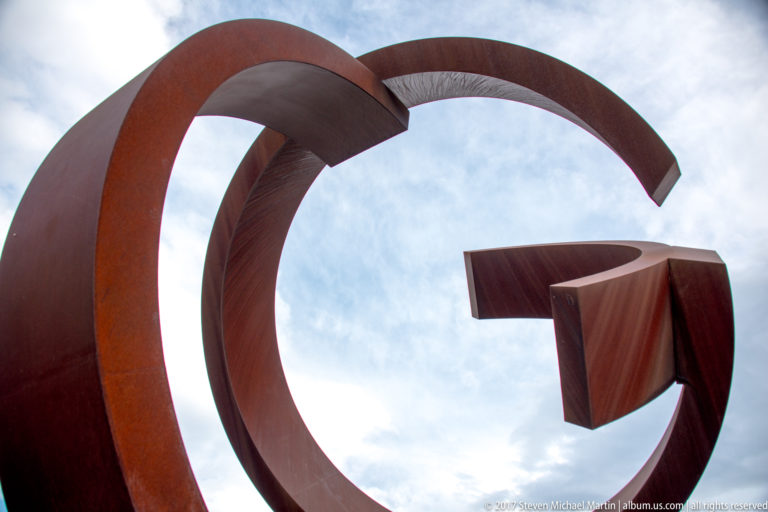 Low-angle view of Jorge Oteiza’s rust-colored abstract steel sculpture formed by sweeping arcs and angular open shapes.