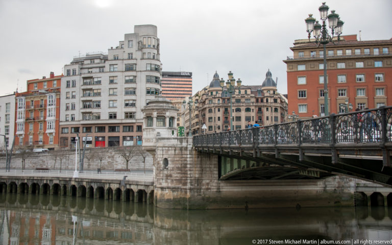 Iron-railed bridge over the Nervión with dense historic and modern city buildings rising along the opposite riverbank.