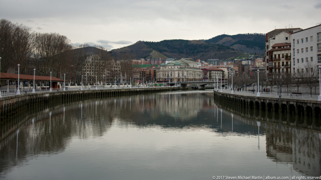Calm Nervión River curving between embankments and city buildings, with wooded hills rising behind central Bilbao.