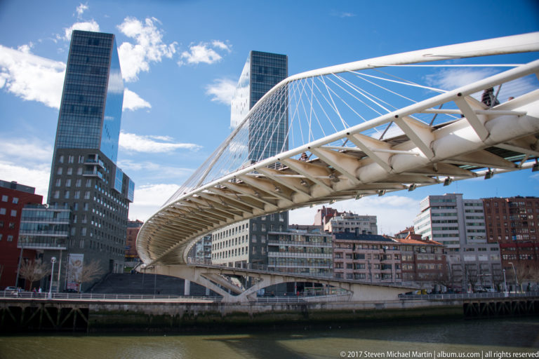 White arched Zubizuri pedestrian bridge crossing the river with a tall modern tower behind it.