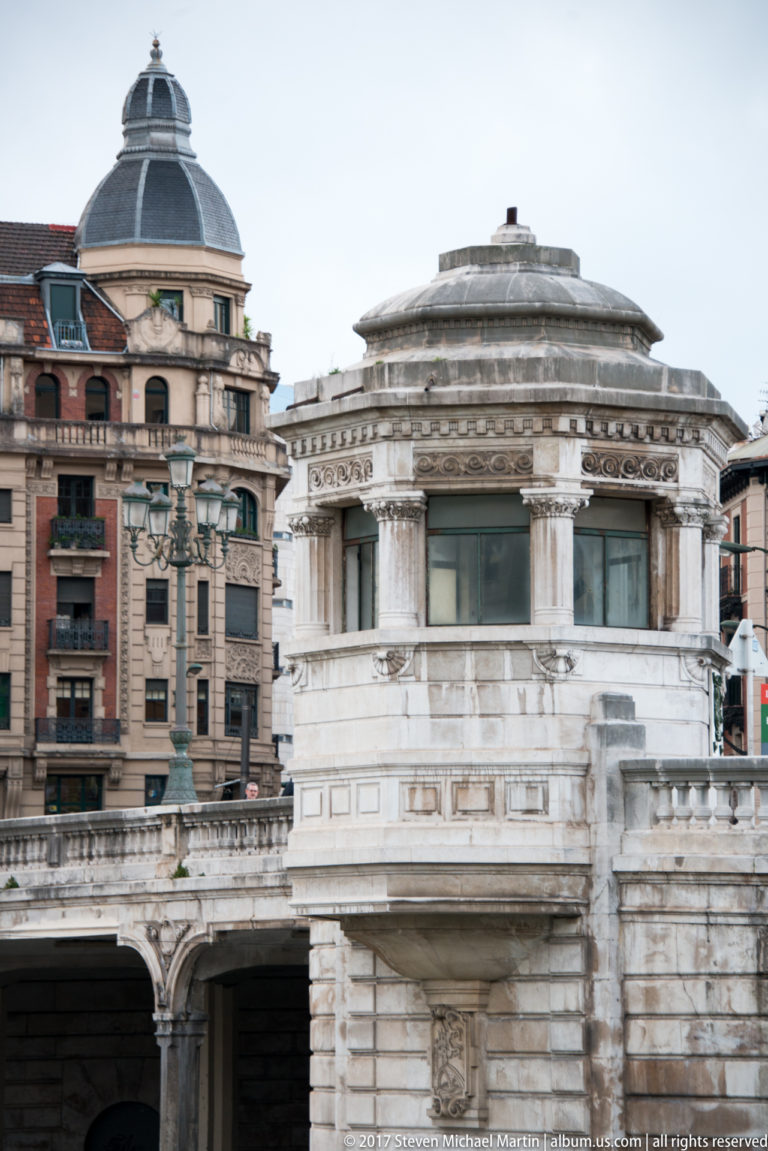 Ornate pale-stone bridge pavilion with circular metal roof, classical detailing, and historic buildings behind it.