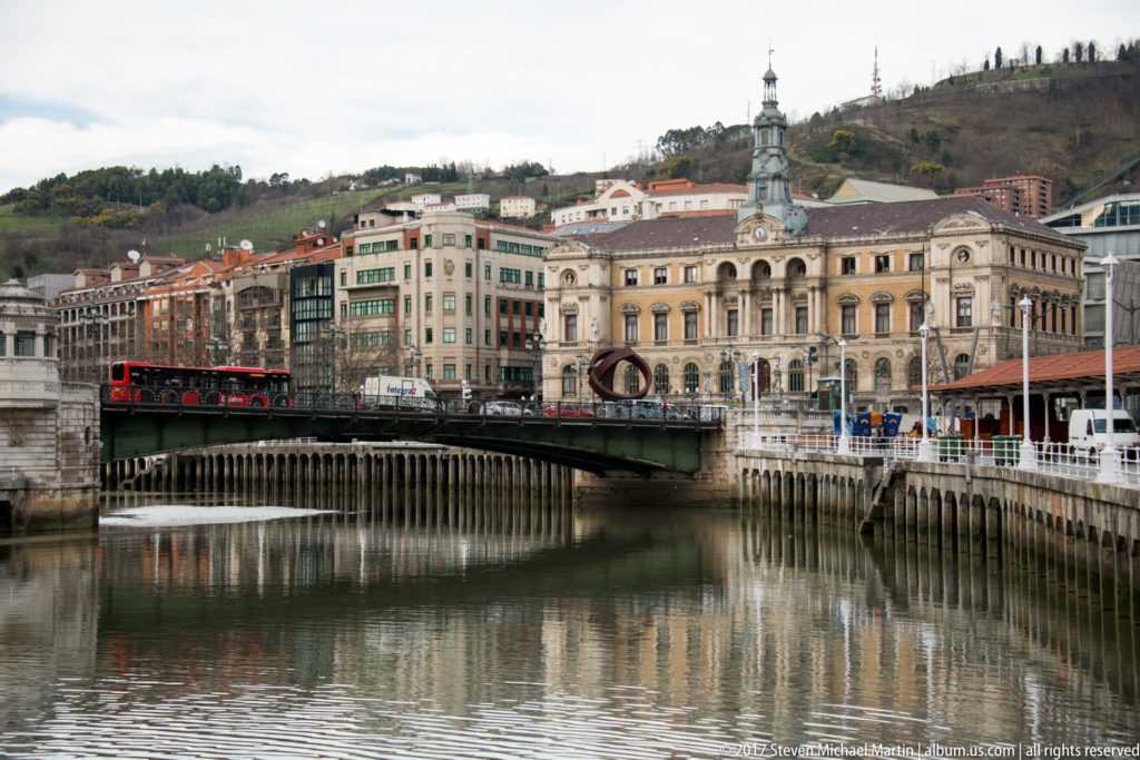 Bridge over the Bilbao estuary beside an ornate historic building, with hillside neighborhoods rising in the background.