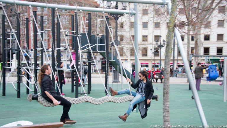 Two adults sit on large rope swings in a public playground surrounded by climbing frames, trees, and pedestrians.