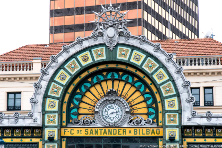 Close view of Bilbao-Concordia station’s colorful arched window, central clock, sculpted surround, and Santander-to-Bilbao railway inscription.