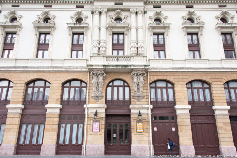 Lower façade of Teatro Arriaga with tall arched doors, balconies, pale stonework, and paired sculptural architectural figures.