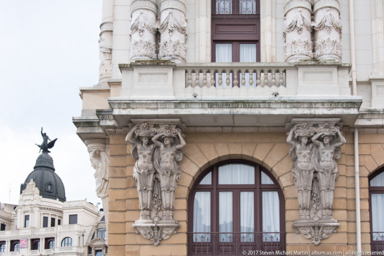 Teatro Arriaga façade detail with paired carved figures around an arched window and a neighboring domed rooftop at left.