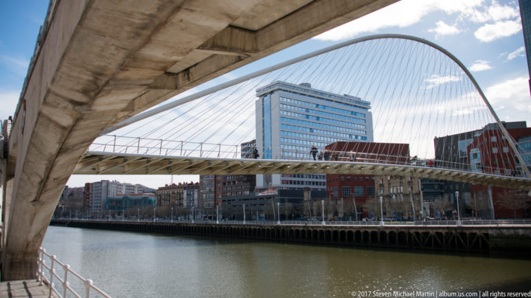 Zubizuri’s white arch and cables seen across the river from beneath a concrete bridge.