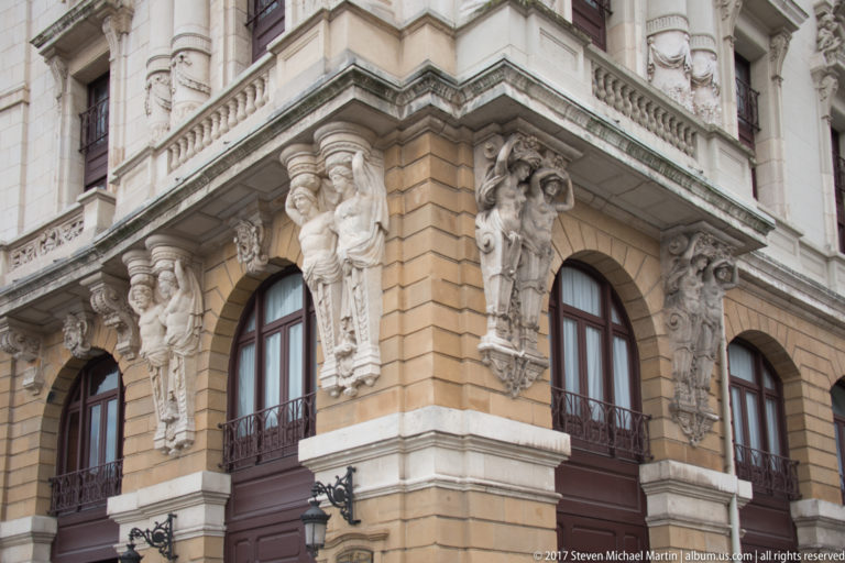 Angled Teatro Arriaga corner with carved human figures, arched windows, balconies, and rusticated tan stone walls.