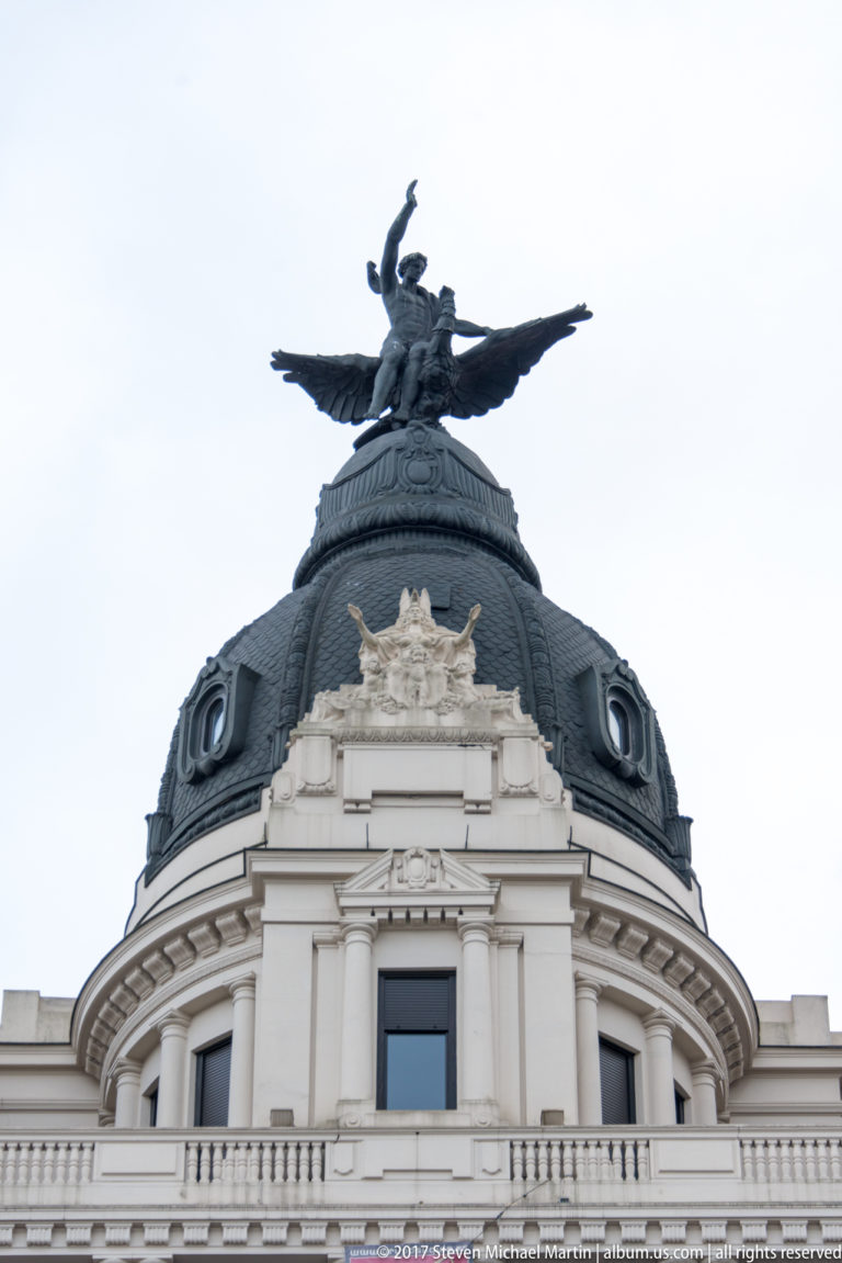 Winged sculptural figure crowns a dark rounded dome with dormer windows above a pale classical building façade.