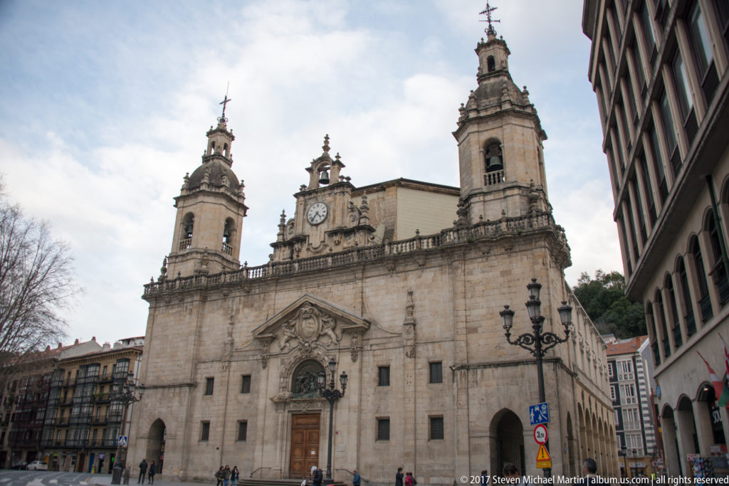Stone façade of the Church of Santos Juanes with twin bell towers, central clock, carved portal, and pedestrians below.
