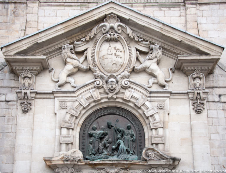 Church portal detail with two carved lions supporting a shield above a round bronze relief filled with religious figures.