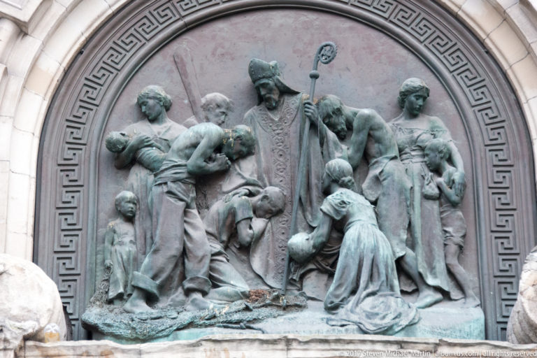 Round bronze relief with a mitred cleric holding a crozier, surrounded by kneeling women and children.