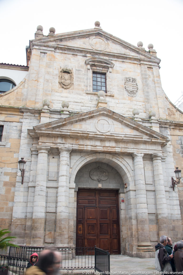 Weathered stone church façade with a large wooden door, paired columns, classical pediment, and small upper windows.