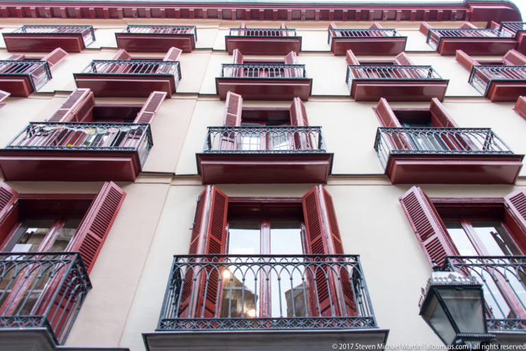 Upward view of a pale residential façade with repeated wrought-iron balconies, red shutters, and tall windows.