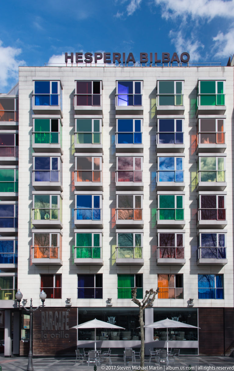 Hesperia Bilbao façade with rows of projecting windows screened by blue, green, orange, and purple glass panels.