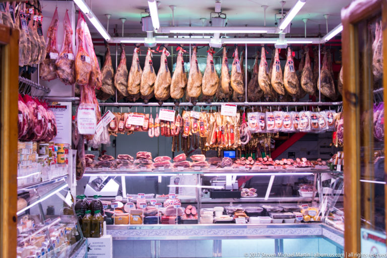 Rows of cured hams hang above a refrigerated meat counter filled with sausages and sliced products in a Bilbao shop.