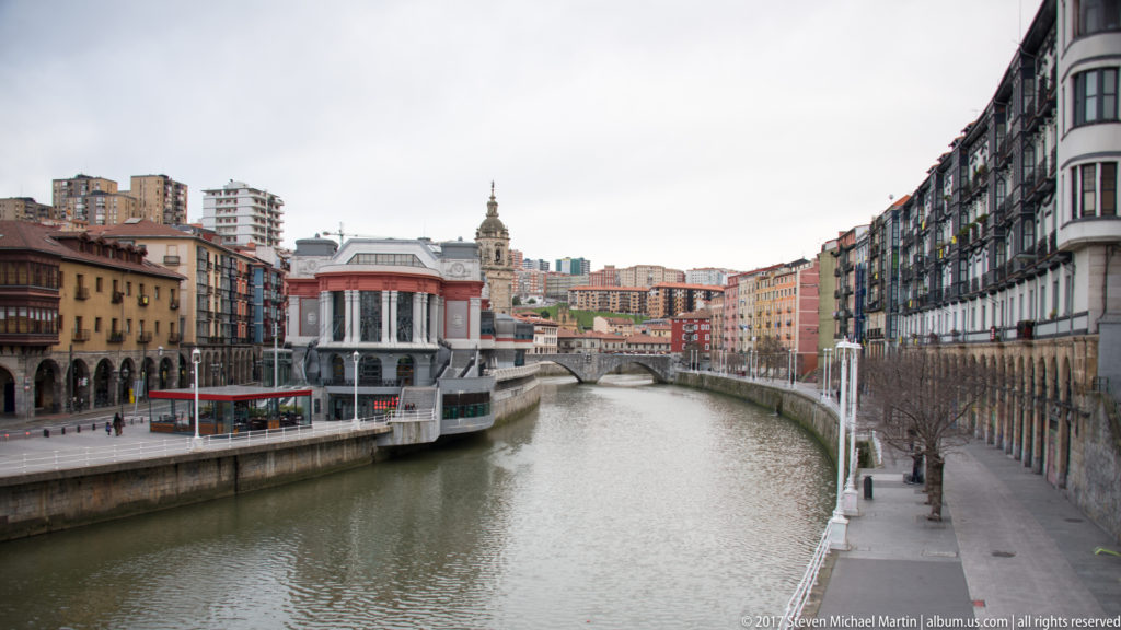 Bilbao estuary between dense rows of buildings, with quays, a church tower, and hillside apartment blocks in the distance.