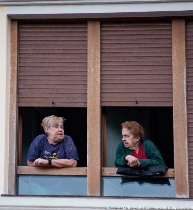 Two women lean from adjacent open windows beneath brown shutters, facing one another across a narrow wall division.