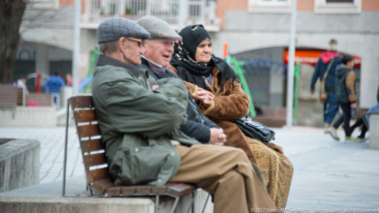 Three older adults in coats and hats sit talking on a wooden bench in a Bilbao public square.