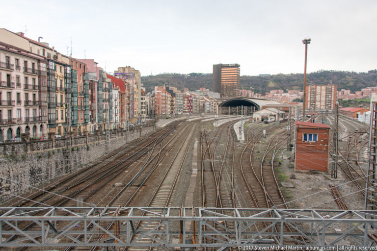 Multiple railway tracks, platforms, and rail equipment run between dense Bilbao apartment blocks toward wooded hills.