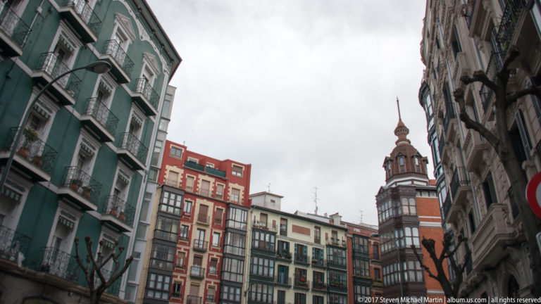 Narrow Bilbao street framed by tall apartment façades, balconies, bare trees, and a red-and-stone tower in the distance.
