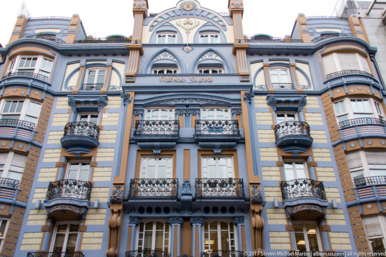 Blue-and-ochre historic façade with stacked wrought-iron balconies, curved bays, arched windows, and decorative roofline details.