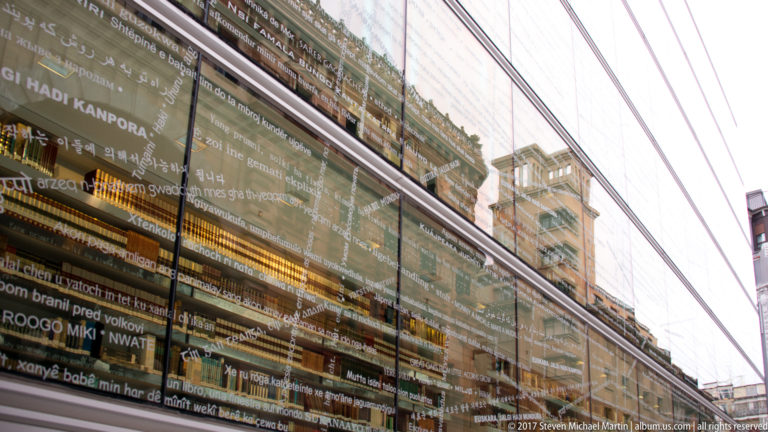 Oblique glass façade at Azkuna Zentroa covered with multilingual text and layered reflections of surrounding Bilbao buildings.