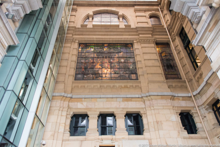 Large warm-toned figurative stained-glass panel set high in a pale stone interior wall above a row of dark windows.