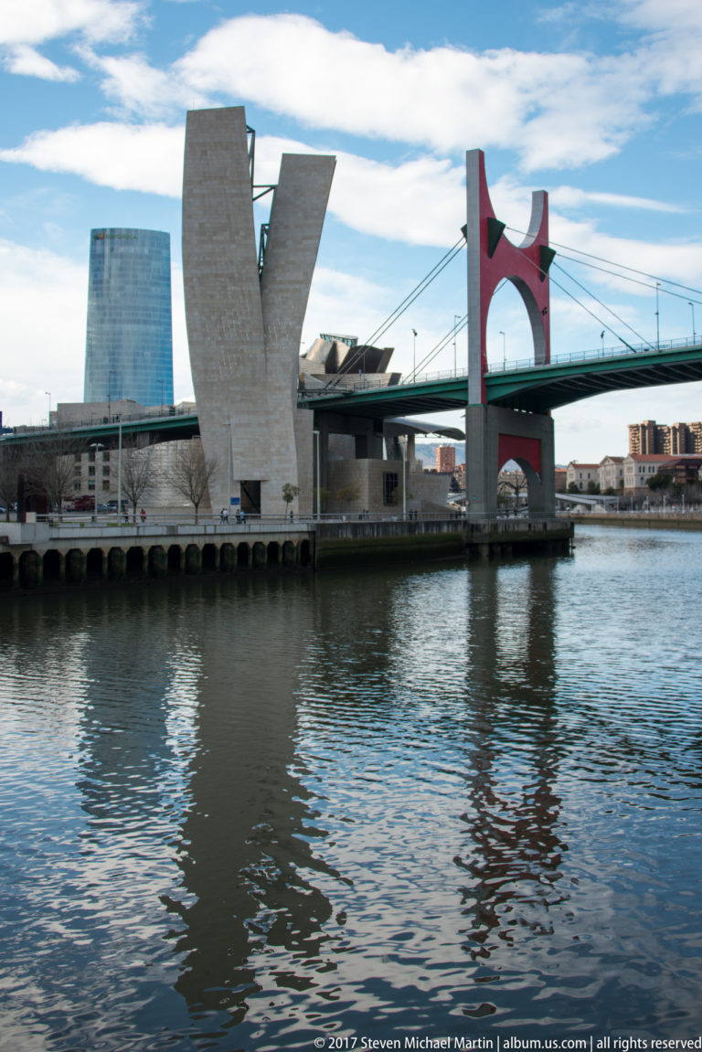 La Salve Bridge with red arch beside pale angular Guggenheim Museum forms across the Nervión River.