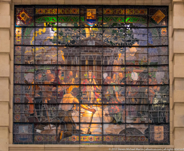 Large multicolored stained-glass window with many figures gathered around a bright central scene, framed by decorative borders and heraldic motifs.
