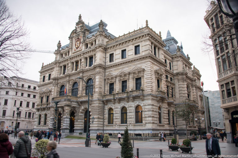 Grand stone provincial government building with arched windows, balconies, sculpted roofline, crest, and pedestrians at a Bilbao intersection.