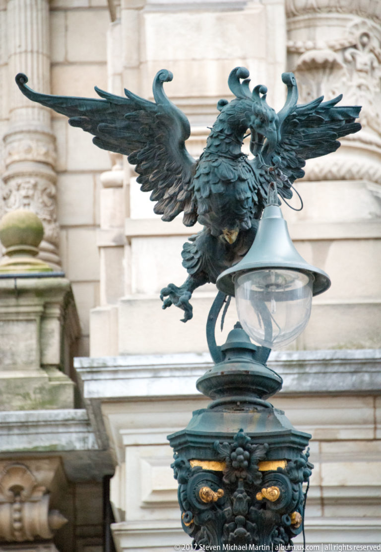 Patinated metal eagle with spread wings perched above a glass globe and ornate dark metal base against a stone building.