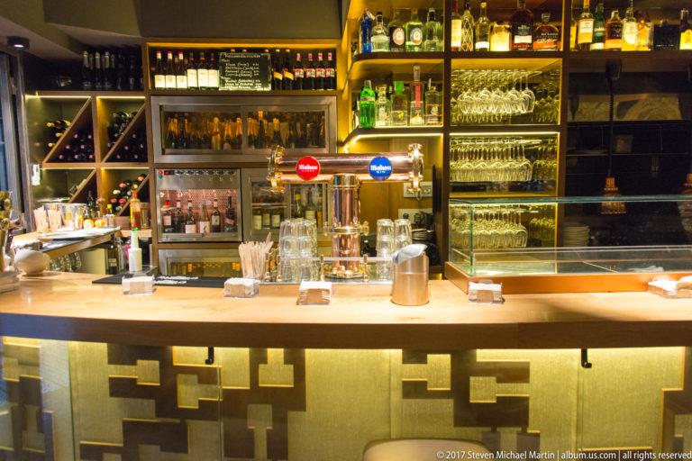 Warmly lit restaurant bar with bottles, hanging wine glasses, beer taps, coffee equipment, and a metallic geometric counter front.