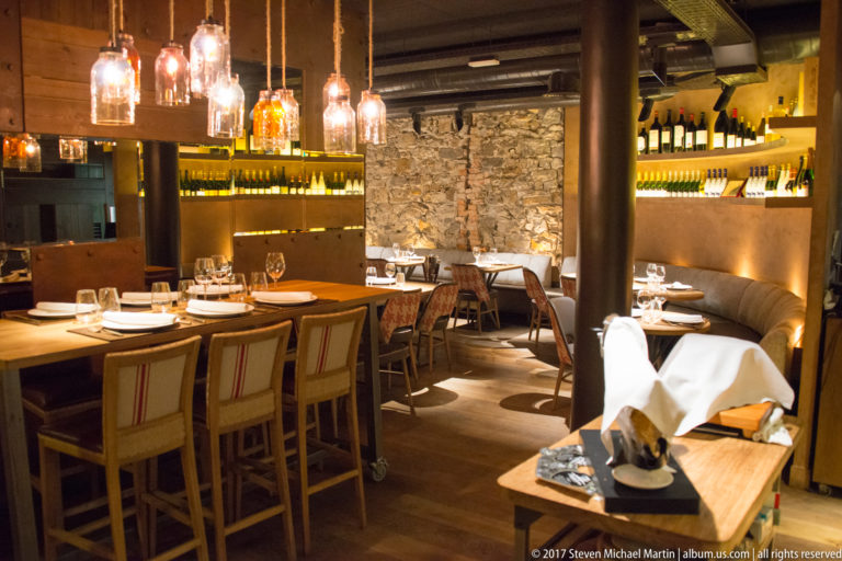 Cozy restaurant dining room with set tables, pendant lights, exposed stone wall, wine shelving, and upholstered seating.